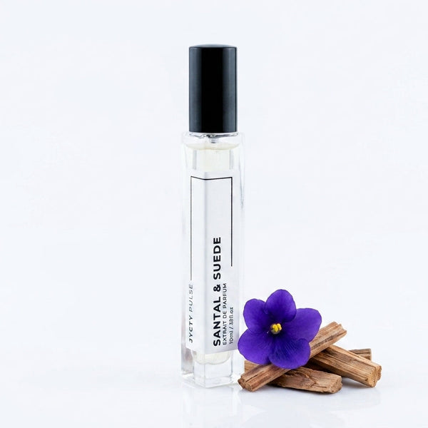 A clear glass perfume roller bottle with a black cap and white label reading 'SANTAL & SUEDE' by JYCTY, styled with a purple flower and cinnamon sticks on a white background.