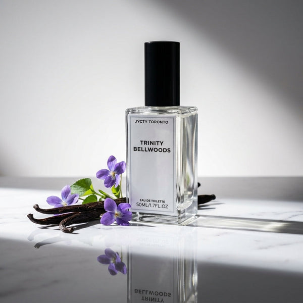 A clear 50ml glass perfume bottle labeled 'TRINITY BELLWOODS' with a black cap, set on a reflective white marble surface beside purple flowers and vanilla beans against a gray background with dramatic shadows.
