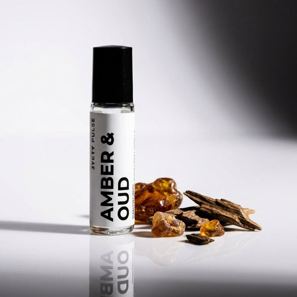 A clear glass rollerball bottle labeled 'AMBER & OUD' with a black cap stands on a reflective white surface beside amber resin pieces and dried bark pieces. The background is a clean white studio setting with a dark shadow on the right.