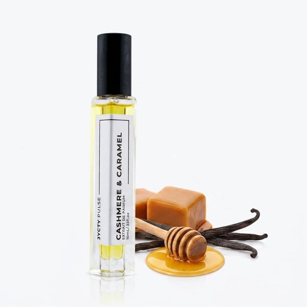 A clear cylindrical perfume roller bottle with a black cap and white label reading 'CASHMERE & CARAMEL' BY JYCTY', shown on a white background with caramel, vanilla beans, and a honey dipper as flavor props.
