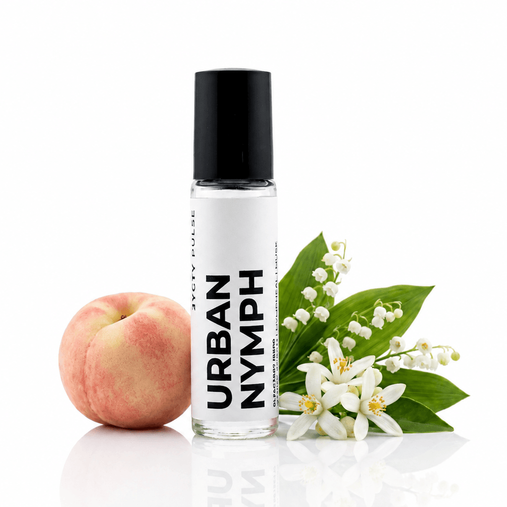 A clear glass roller bottle with a black cap labeled 'URBAN NYMPH' stands on a white reflective surface beside a peach and white flowers with green leaves. The brand 'JYCTY' is visible on the bottle.