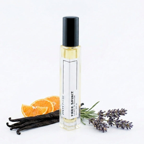 A clear glass roller bottle with a black cap and white label reading 'JYCTY PULSE FREE SPIRIT' stands on a white background, styled with vanilla beans, orange slices, and lavender sprigs.