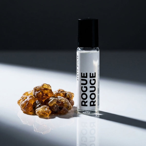 A clear cylindrical perfume oil bottle labeled 'ROGUE ROUGE' with a black cap stands on a white reflective surface beside several amber-brown resin-like pieces against a dark gray background.