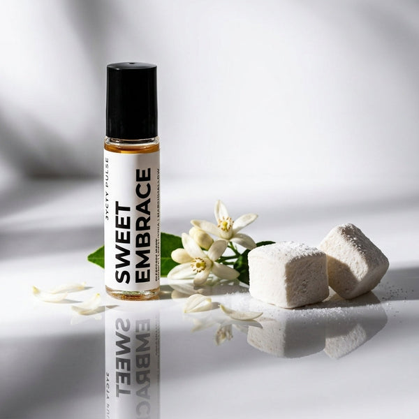 A small roll-on bottle labeled SWEET EMBRACE stands on a glossy white surface with white flower petals, white sugar cubes, and a few green leaves around it. The background is a softly lit white studio setting with dramatic shadows.