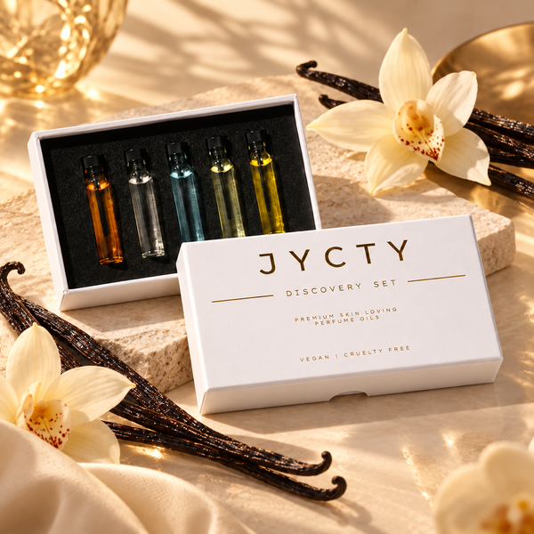 A boxed fragrance discovery set on a warm beige fabric surface with flowers and vanilla beans. The set includes a white box labeled 'JYCTY DISCOVERY SET' and an open tray showing five small perfume oil vials with amber, clear, blue, yellow, and green oils.