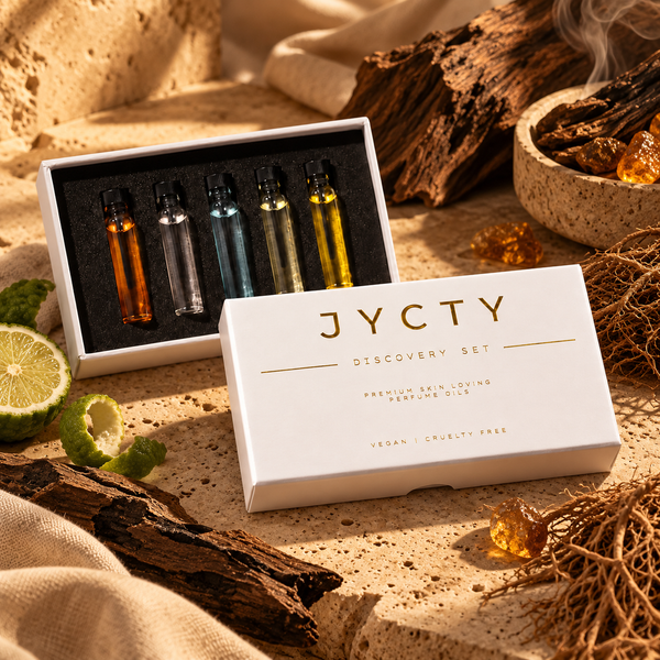 A boxed fragrance discovery set on a warm sandy surface with dried wood, citrus, and amber props. The open white box shows five small perfume oil vials in amber, clear, blue, and yellow tones, with the front lid reading 'JYCTY DISCOVERY SET'.