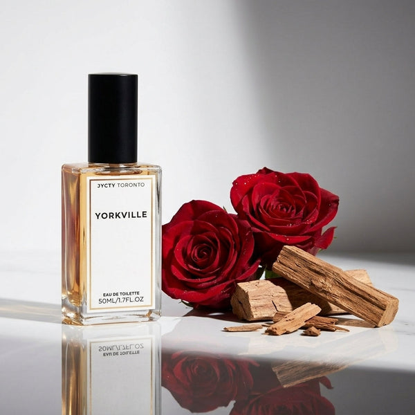 A 50ml bottle of Yorkville Eau de Toilette stands on a reflective white surface beside two red roses and pieces of wood, against a clean light gray background.